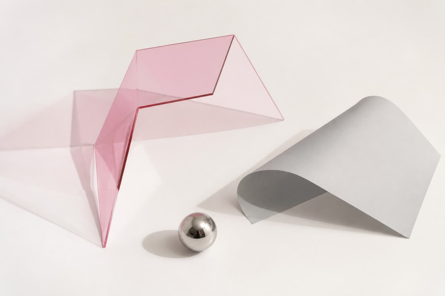 Pink acrylic, folded paper, and a chrome sphere arranged on a white surface